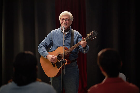 Elderly Caucasian man performing on stage playing acoustic guitar and singing into microphone during talent competition smiling at audience with people watching in foreground - Powered by Adobe