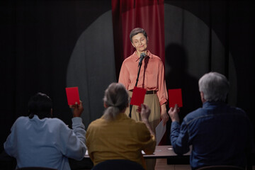 Caucasian woman standing on stage singing into microphone during talent competition while three judges holding up red cards in foreground