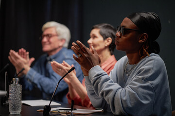 Three judges sitting at table clapping hands during talent competition, including senior Caucasian man, middle aged Caucasian woman, young adult Black man wearing sunglasses