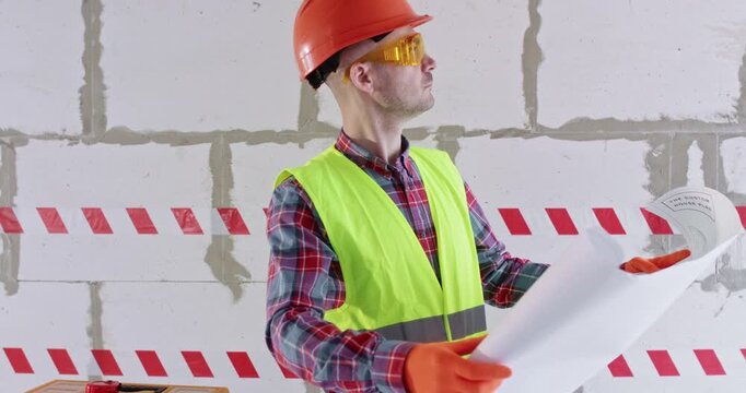 Man in safety glasses examines floor plan standing in hall. Bearded guy studies apartment layout before starting renovations and analyzes future scope of work
