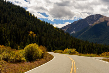 Naklejka premium In the center of the road, surrounded by towering pines, with a mountain peak ahead under a dramatic, cloud-filled sky.