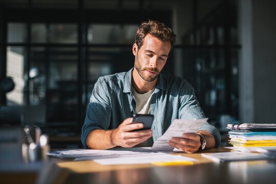 Man scanning paper receipts with smartphone app at office desk, integrating paperless technology into modern bookkeeping for improved expense tracking, data organization, and financial efficiency.