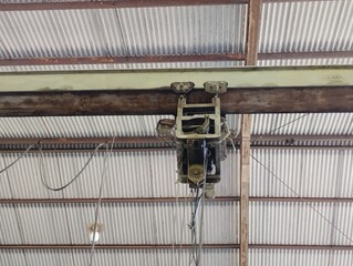 Close-up view of an industrial overhead crane hoist mechanism mounted on a steel beam inside a warehouse. The equipment shows signs of use and aging, with visible rust and electrical cables hanging