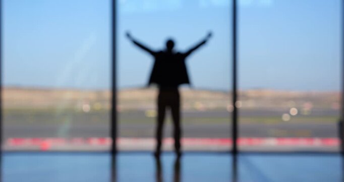 In airport terminal, man in casual jacket celebrates, raising his hands in excitement, his figure shown blurred in slow motion. Joyful silhouette, seen from behind, captures thrill of success