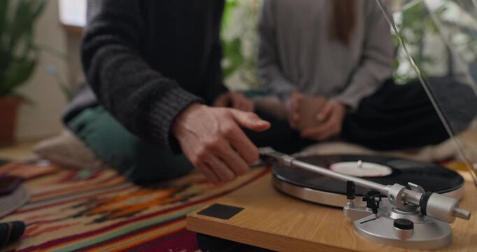 Close up of a guy putting a vinyl record in a player and turning it on while listening to vintage music with his girlfriend and having tea at home in a cozy apartment