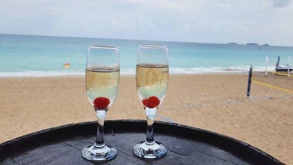 Champagne flutes with cherries stand on a barrel top, overlooking a beautiful tropical beach with sea shore. travel and summer holidays trip concept. 4k video. - Powered by Adobe