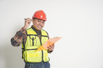 Asian construction worker wearing safety vest and helmet, holding a clipboard while pointing and writing. Isolated image on white background
