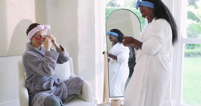 Diverse female friends entering lounge, exchanging pink under-eye patches by mirror for skin care