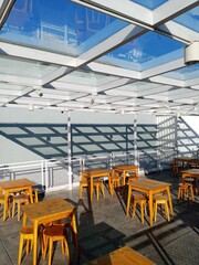 Description: An outdoor seating area with a modern glass and white metal framed roof, casting geometric shadows onto the wall and floor. Wooden tables and stools are visible underneath the structure