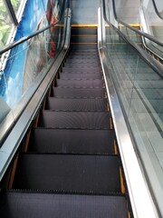 Obraz premium A high-angle view of a modern escalator with black treads and a glass balustrade, ascending to a higher floor. The side features a large advertising poster