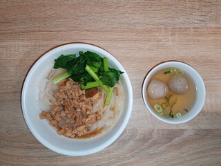 A top-down shot of a bowl of noodles with minced chicken and vegetables, served with a side bowl of bakso meatballs in clear broth
