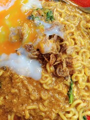 A close-up of a bowl of instant noodles with a rich, savory sauce, topped with a runny fried egg and crispy fried shallots