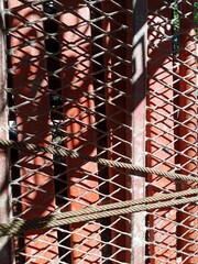 A close-up, high-angle shot of a diamond-shaped metal mesh and thick rope with a red brick wall background, creating strong shadows from the sunlight
