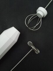 A simple, minimalist flat lay of a battery-operated milk frother with its two different whisk attachments, on a dark, plain background