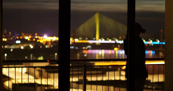 Silhouette of businessman pacing on balcony, talking on phone at night. City lights and illuminated bridge visible blurred in background. Urban lifestyle, communication at nighttime