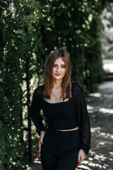 Girl in black trousers and a black blouse against the background of green leaves in summer she has long hair