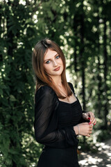 Girl in black trousers and a black blouse against the background of green leaves in summer she has long hair