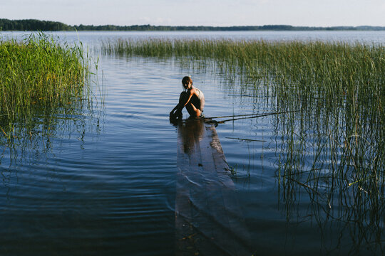 Woman sitting on wooden pier, calm lake waters surrounded by grass