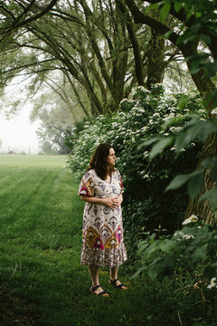 Pregnant woman stands in green trees and fields in floral dress