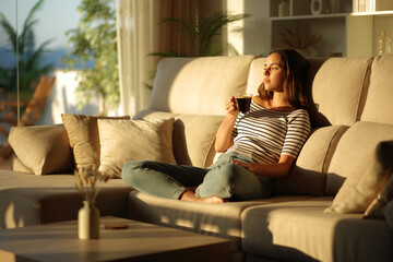 Woman drinking coffee at sunset relaxing on a couch at home