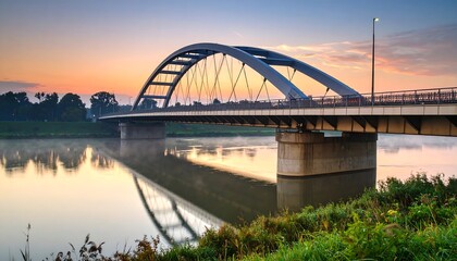 Obraz premium A modern arch bridge spans a tranquil river at dawn