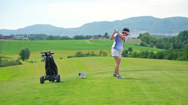 Active senior woman playing golf on a scenic course with rolling hills, enjoying outdoor exercise and vibrant retirement on a sunny summer day.