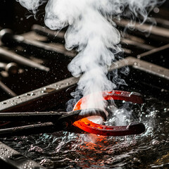 A blacksmith's tongs holding a glowing, red-hot horseshoe, which is being plunged into a tub of water, creating a dramatic burst of white steam and a splash of water droplets