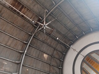A view of the metal arched ceiling of a building interior, featuring a large, industrial-style fan hanging from the center and a round, stained-glass window