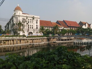 A scenic shot of historical Dutch colonial-style buildings along the Kali Besar river in Jakarta's Old Town, with the water in the foreground and a hazy sky above