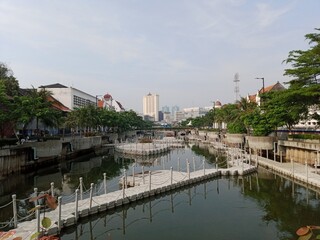 A panoramic shot of the Kali Besar river in Jakarta's Old Town, with the water flanked by walkways, floating platforms, and historical Dutch colonial-style buildings under a hazy sky