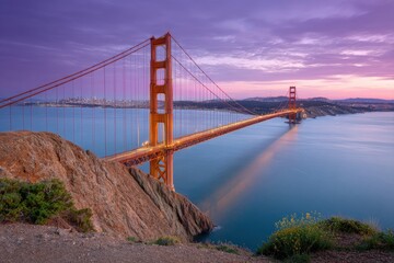 Fototapeta premium Golden Gate Bridge spans blue water under a pink and purple sunset sky with rocky cliffs in the foreground
