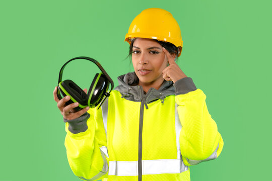 Construction worker wearing a hard hat and safety jacket pointing at her head while holding headphones in front of a green background