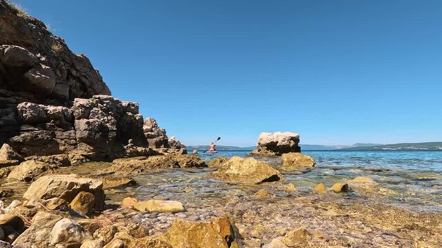cenic Summer View of FKK Konobe Rocky Beach near Punat, Krk Island, Croatia Adriatic Sea, Coastal Cliffs, Islands and Naturist Woman Paddleboarding in Distance under Clear Blue Mediterranean Sky