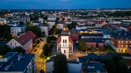 Evening aerial view of Ostrów Wielkopolski featuring the illuminated Church of the Blessed Virgin Mary and surrounding residential buildings.