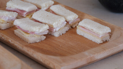 Delicate ham and cheese finger sandwiches layered with creamy sauce resting on rustic wooden serving board, presenting elegant party appetizer