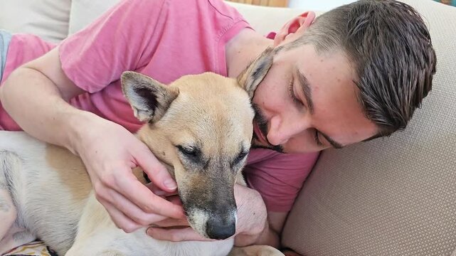 White man lying on sofa cuddling mixed breed dog with short light brown fur and black muzzle while gently holding its head and resting face against pet in home interior close up emotional moment