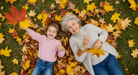 Senior grandmother and smiling granddaughter lying on yellow and red leaves at park, view from above. Top view of playful old woman with cheerful girl lying on autumn park leaves, looking at camera.