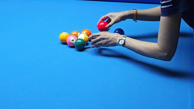 Hands arranging colorful billiard balls into triangle rack formation on blue pool table surface before game start close up of player preparing balls for break shot in indoor game setting