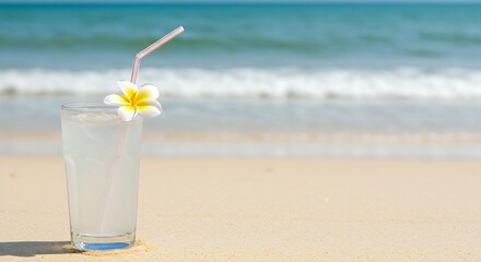 A glass of pure coconut water with a straw, garnished with a tropical flower, on a sandy beach background with blurred ocean waves. 