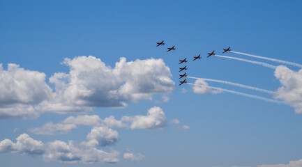 squadron of nine jet planes performs an aerial formation against a bright blue sky, leaving white trails behind