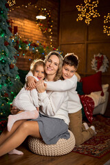 smiling mother and her two children in white sweaters hug closely, celebrating in a cozy, festive room with a decorated tree and golden lights