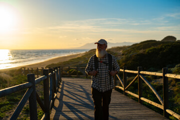 Fototapeta premium Senior Man with a White Beard Walking Along a Scenic Coastal Boardwalk at Sunset, Wearing a Plaid Shirt and a Backpack, Enjoying the Tranquility of Nature
