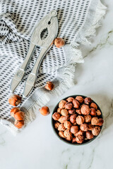 Minimal rustic composition — metal nutcracker resting on woven cotton cloth next to bowl of dried hazelnuts, shot in soft daylight on marble background with negative space