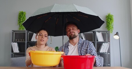Frustrated couple sitting indoors under an umbrella with buckets collecting water from a leaking ceiling. Concept of housing issues, apartment maintenance, and home repair problems. 