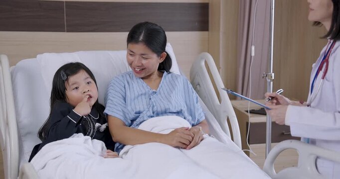 asian female patient lying on hospital bed smiling with young asian female child sitting beside during heartwarming family visit while doctor stands nearby preparing for medical consultation and care