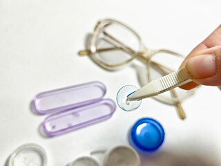 Careful handling of contact lens with tweezers near eyeglasses and storage cases on white surface