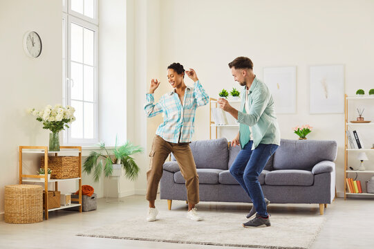 A cheerful young couple dancing together at home in a bright modern living room. The image conveys joy, relaxation, and positive emotions in a domestic lifestyle setting. 