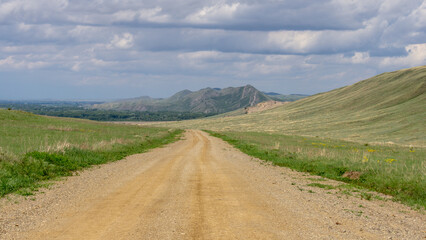 Long mountains. Scenic dirt road winding through lush green hills under a cloudy sky, leading towards distant mountains, inviting exploration and adventure in a serene natural landscape