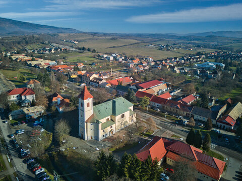 Aerial view of a church with green roofs and a red-tipped tower stands amidst a village nestled in a valley, its buildings casting long shadows, Detva, Bansk&Atilde;&iexcl; Bystrica Region, Slovakia.