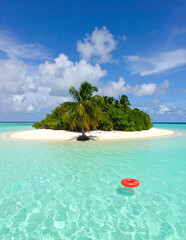 Small isolated sandy island with single palm tree and red inflatable ring in shallow clear turquoise water under bright blue summer sky